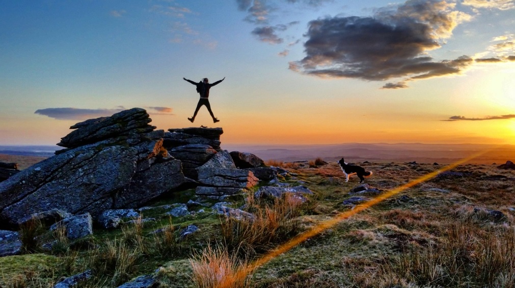 Photo of a walker jumping for joy on rocks at Leeden Tor, Dartmoor at sunset while a dog watches